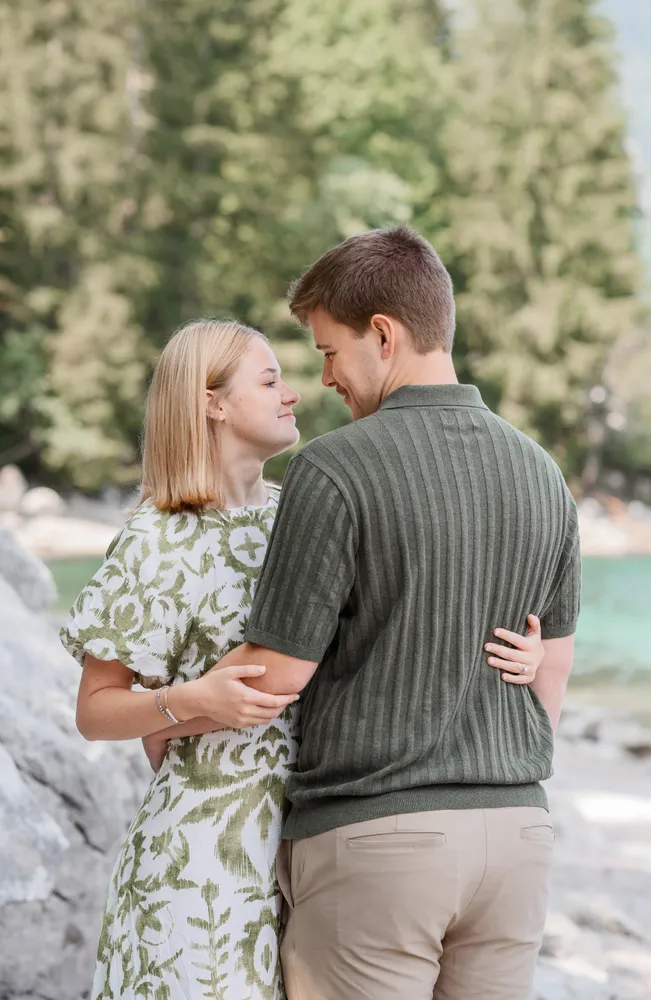Couple in casual attire embracing by a serene lake, surrounded by lush greenery. Their faces are turned towards each other, reflecting a moment of intimacy and affection. The man wears a dark green striped polo and khaki pants, while the woman is dressed in a white dress with a green floral pattern.