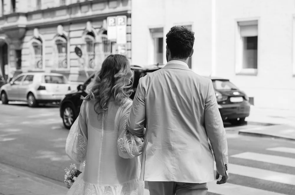 Couple in wedding attire walks down urban street, hand-in-hand, facing away from camera. Black and white photo captures candid moment.