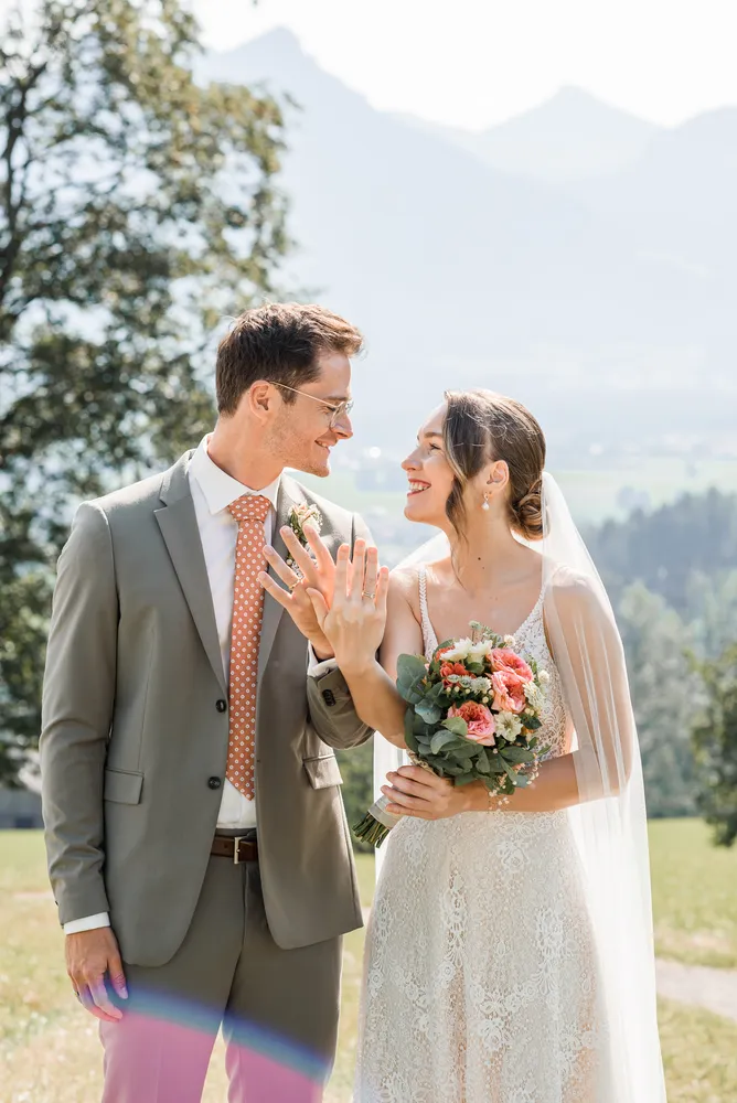 Bride and groom in wedding attire stand outdoors, smiling and holding hands. She wears a white lace dress with veil, holding a bouquet. He is in a grey suit with a patterned tie. Background features blurred natural scenery.