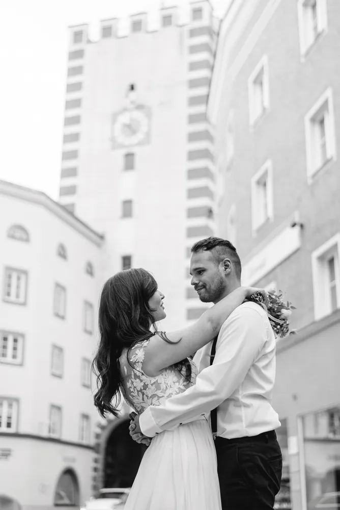 A couple embracing in a city street, the woman in a white lace dress and the man in a white shirt and black pants. They stand before historic buildings with a clock tower in the background.