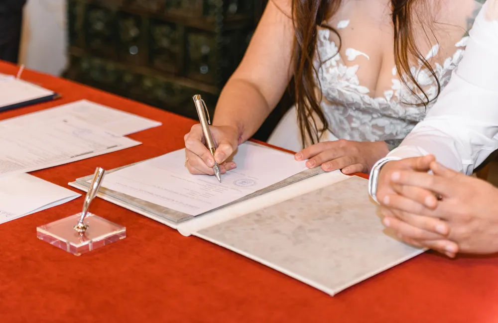 Woman in white lace dress signs document with gold pen at red-covered table, beside man's clasped hands, formal setting.