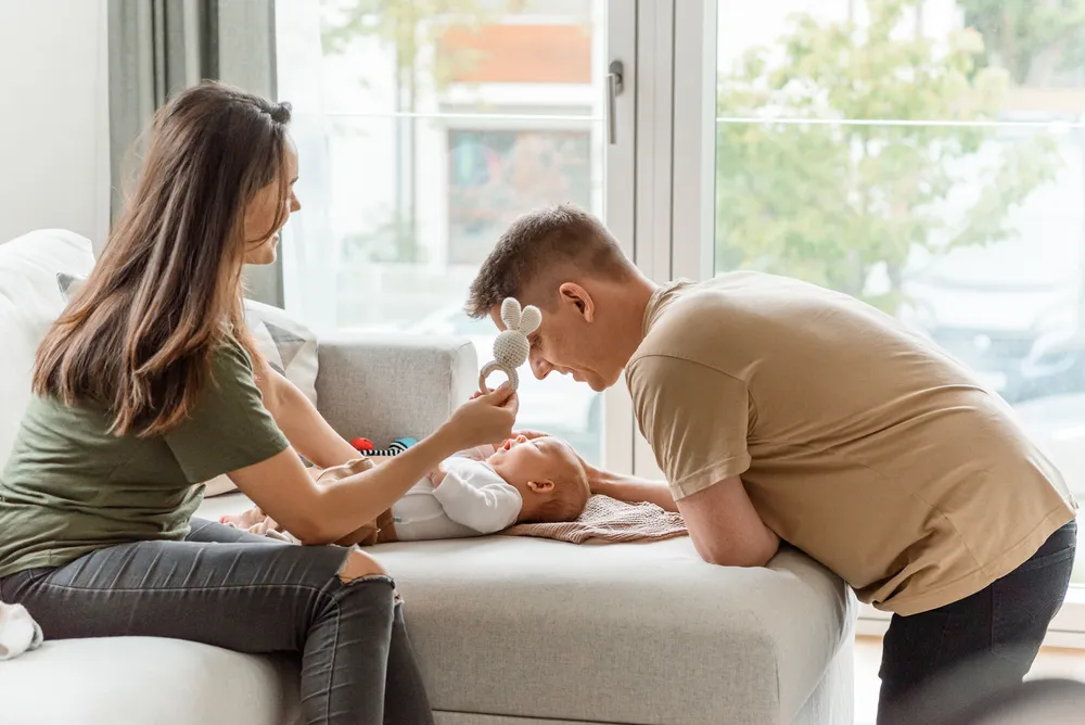 Parents interact with baby on modern sofa. Woman holds bunny toy, man leans in. Large window, natural light, urban backdrop.