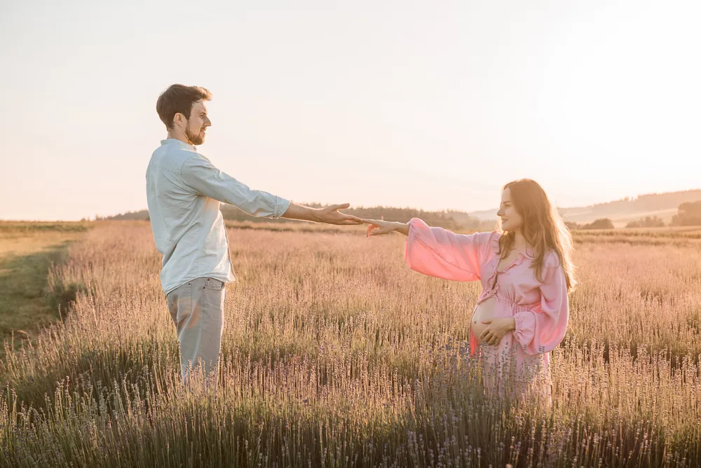 Couple in a lavender field at sunset, woman in pink maternity dress reaches for man's hand, both smiling, serene landscape in background.