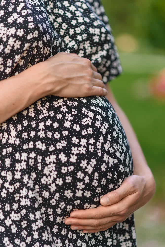 Pregnant woman in a black-and-white floral dress gently cradles her belly outdoors, with a blurred green background.