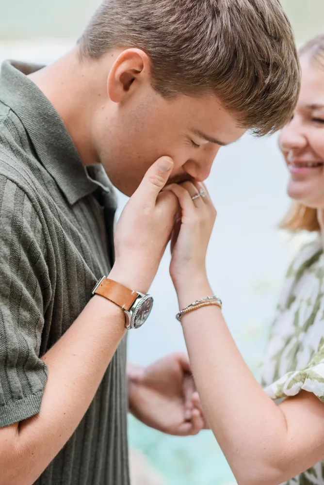 Man in green shirt receives a ring from woman in white dress. They stand close, smiling, outdoors. Focus on their hands and faces.