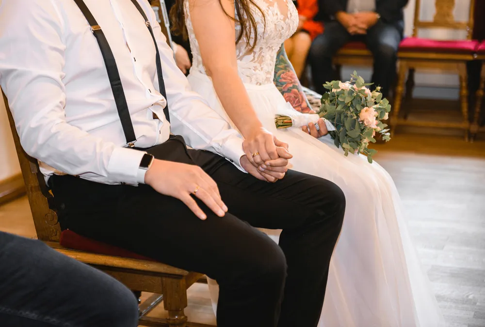 Bride and groom hold hands during wedding ceremony. She wears white lace gown, holds bouquet. He wears white shirt, black trousers, suspenders. They sit on wooden chairs with red cushions. Blurred guests in background. Indoor setting.