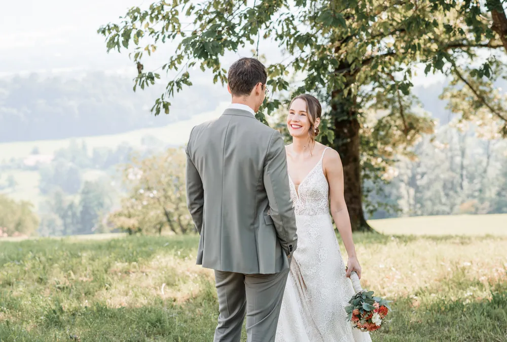 Bride and groom in wedding attire stand in a sunlit meadow under a tree, holding hands and smiling at each other.