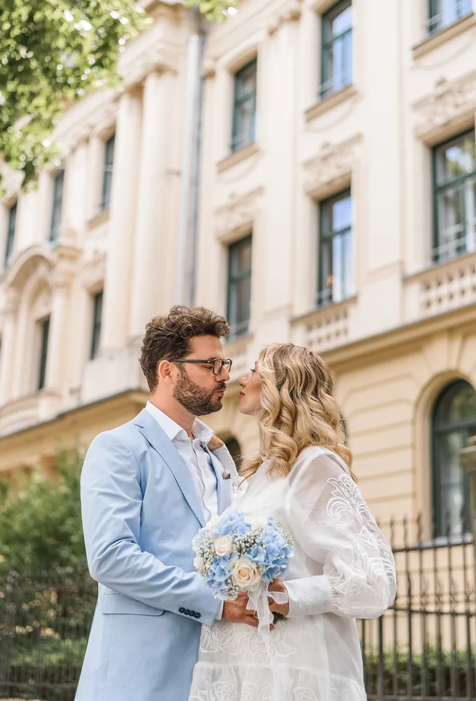 Couple in light blue suit and white lace dress hold hands, standing in front of ornate building with wrought iron fence.