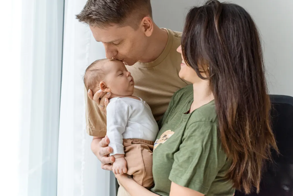Man and woman embrace newborn baby, man kisses child's forehead, woman smiles, domestic setting, natural light.