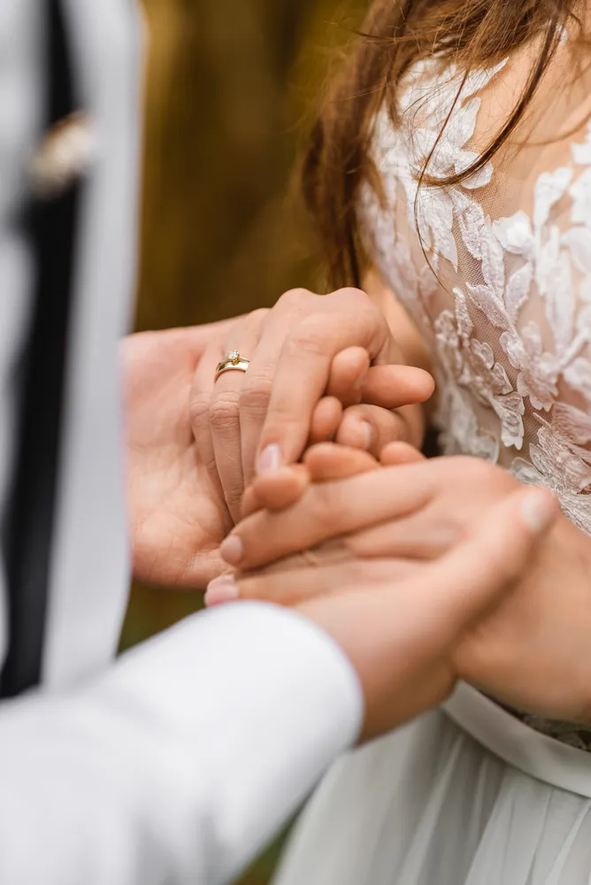 Interlocked hands of a couple, one wearing a ring, against a blurred outdoor backdrop. Formal attire visible. Professional, intimate moment captured.