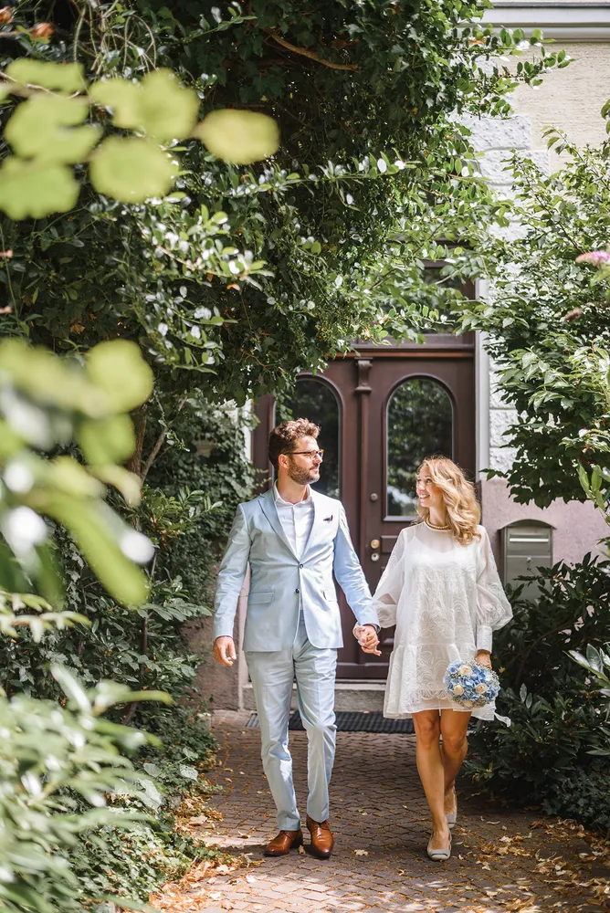 Couple in light blue suit and white dress walks hand-in-hand down a leafy path towards a brown door, holding a bouquet.