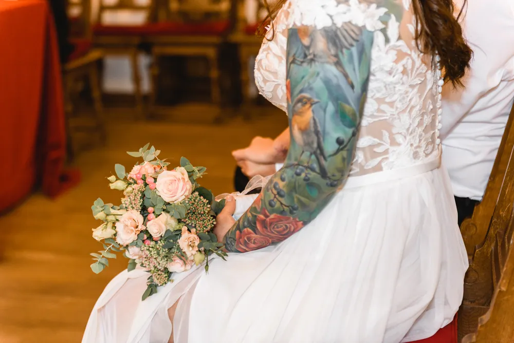 Bride with tattooed arms holds bouquet of pink and white flowers at wedding ceremony, seated beside groom in church.