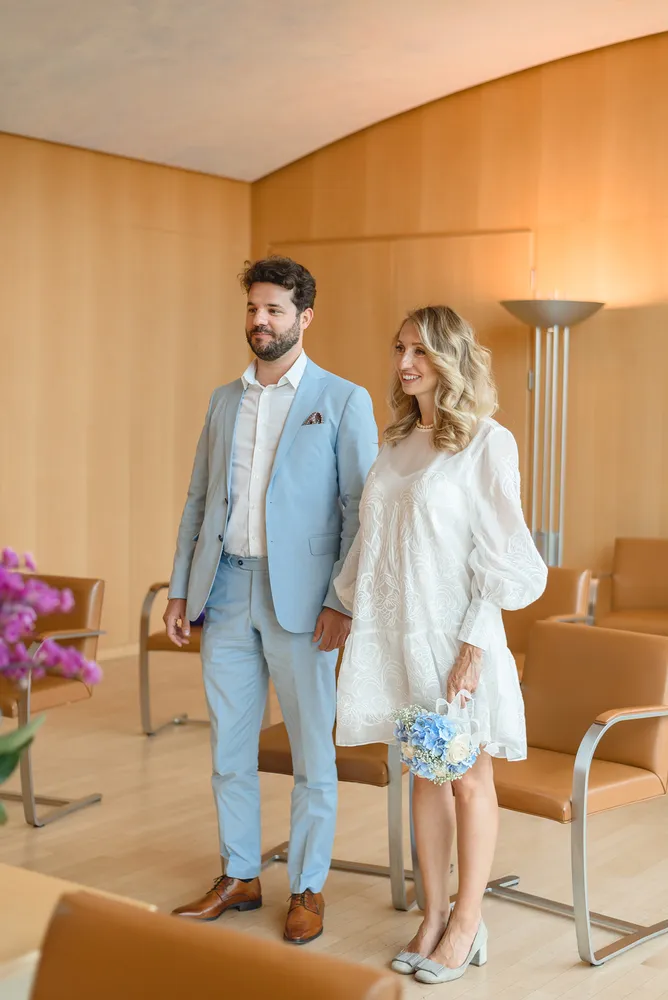 A couple in wedding attire stands in a minimalist room, the man in a light blue suit and the woman in a white lace dress holding a bouquet, surrounded by modern furniture and soft lighting.