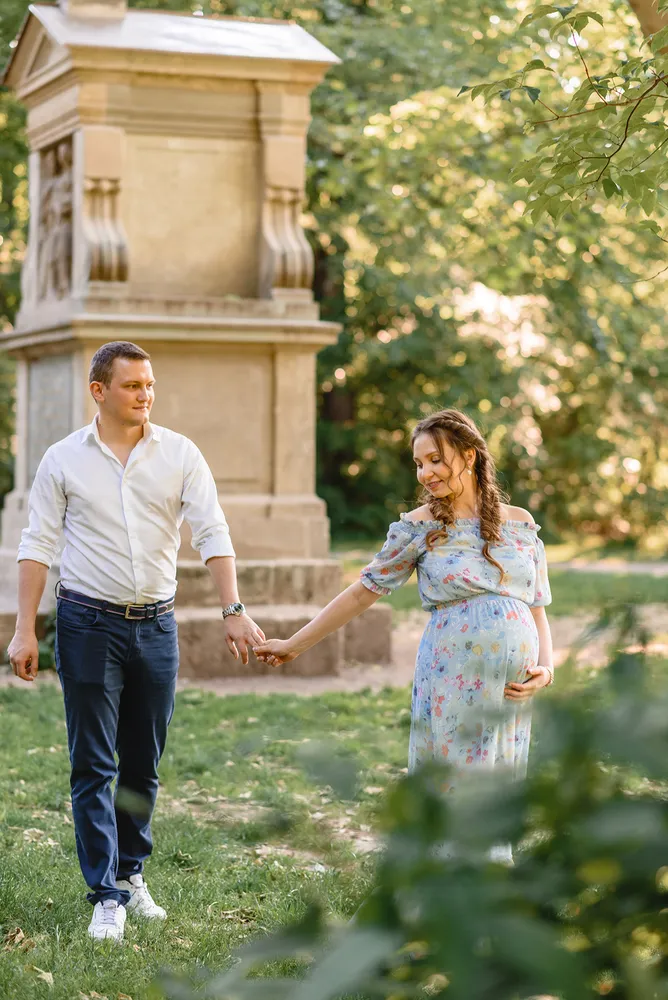 Expectant couple strolls through park, holding hands near historic monument, woman's floral dress reveals pregnancy, serene outdoor setting.