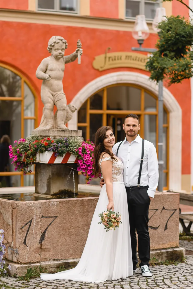 Bride and groom in wedding attire stand by a cobblestone fountain with a cherub statue, vibrant flowers, and a red-orange building with arched windows in the background.
