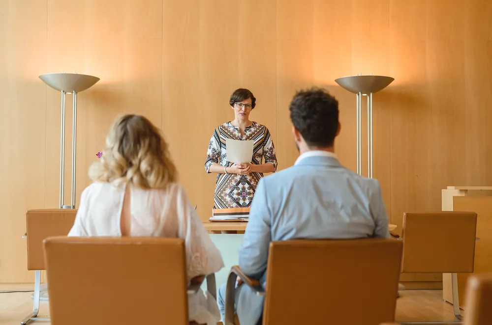 Woman in patterned blouse addresses two attendees in modern conference room with wooden walls and minimalist furniture.