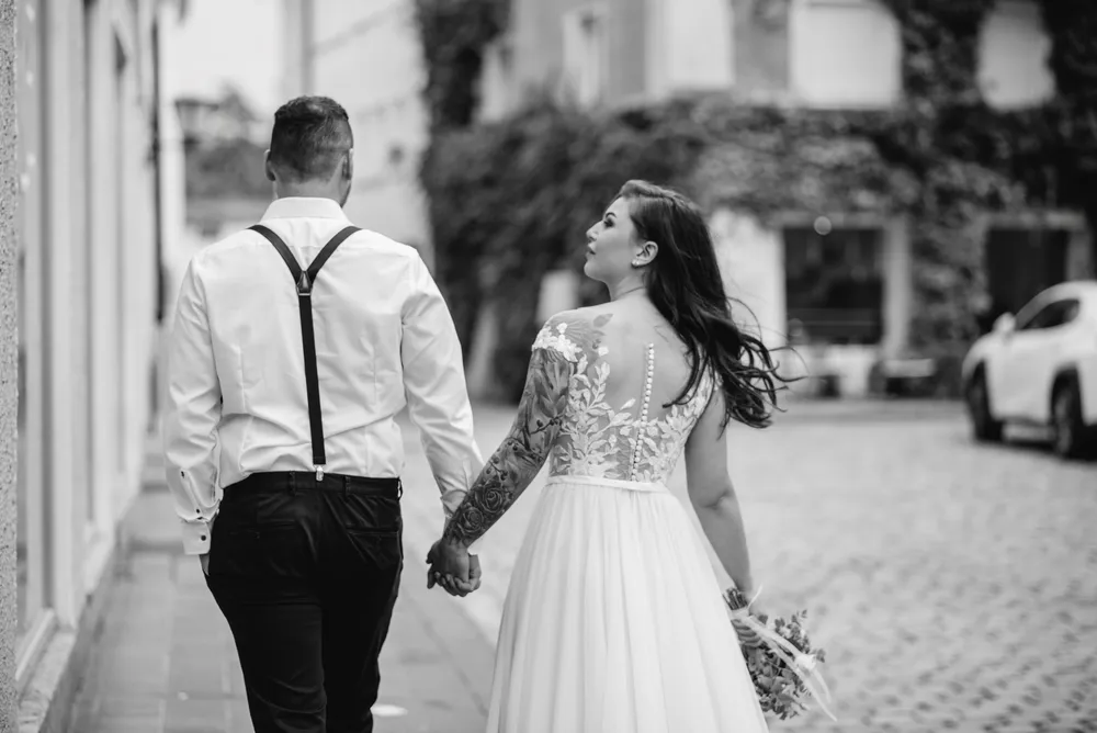 A couple strolls down a cobblestone street, the woman in a white lace wedding dress, the man in a white shirt and black pants. They hold hands, both gazing ahead, with a blurred backdrop of buildings and a car.
