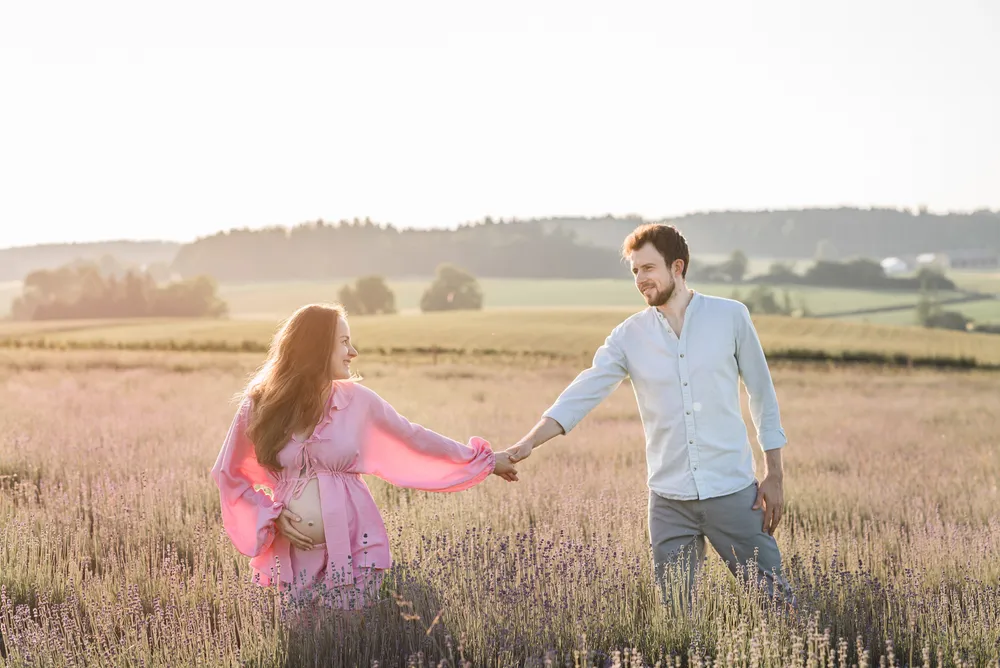 A couple holds hands in a lavender field at sunset, smiling. He wears a light blue shirt and grey pants; she's in a pink dress, cradling her belly. Sunlit, serene landscape.