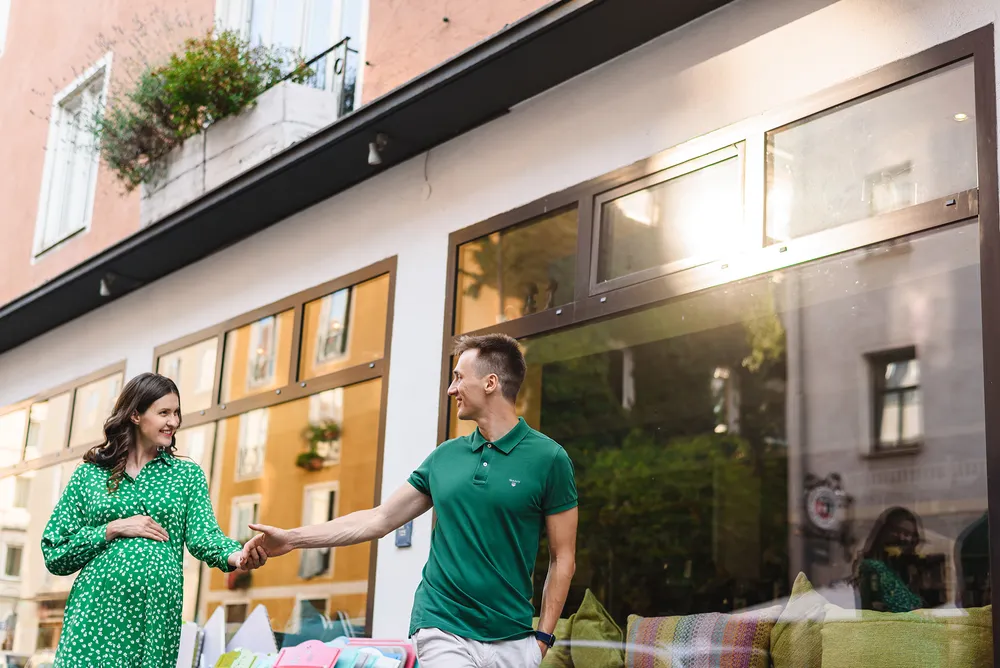 Pregnant woman in green dress shakes hands with man in matching green polo outside modern building with large windows. Sunlit urban street scene.
