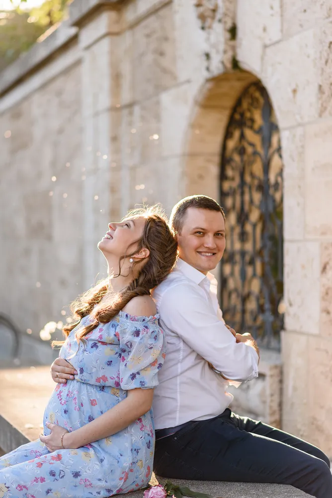 Couple embracing outdoors, woman in floral dress, man in white shirt, seated against stone wall with ornate gate, sunlight filtering through trees.