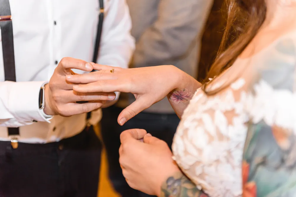 A person in a white shirt with suspenders and a watch assists another individual in placing a ring on their finger, which has a tattoo. The setting appears to be a formal indoor event.
