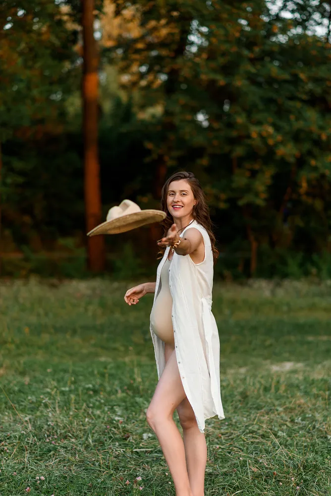 Woman in white dress joyfully tosses wide-brimmed hat in sunlit forest clearing.