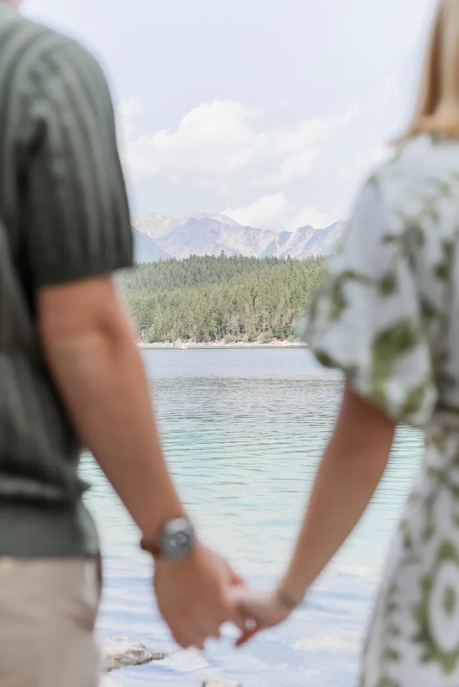 Two individuals, one in a striped shirt and the other in a floral dress, hold hands by a serene lake, with mountains and dense trees in the background, under a partly cloudy sky.