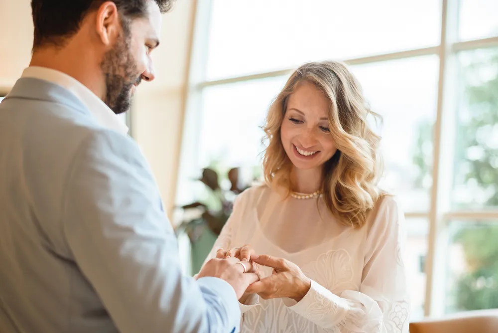 Couple in formal attire exchanging rings indoors, smiling. Natural light through large windows. Blurred foliage visible outside. Professional, intimate moment captured.
