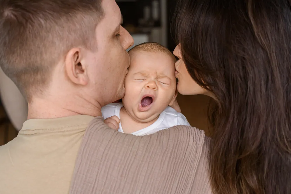 A man and woman kiss a sleeping baby in their arms indoors.