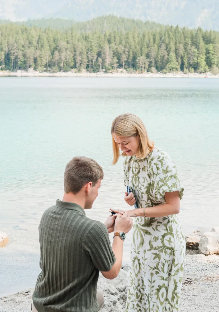 Man proposes to woman at lakeside; she accepts. Scenic backdrop of mountains and forest. Casual attire, natural lighting.