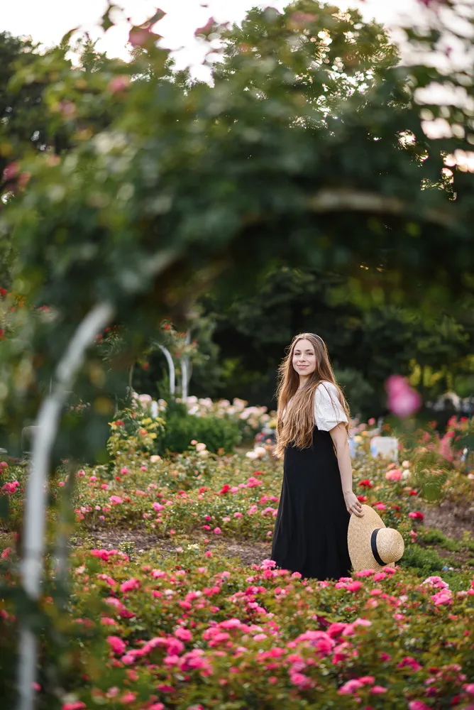 A woman in a black dress stands amidst a vibrant rose garden, holding a straw hat. She is smiling softly, surrounded by lush greenery and blooming flowers, with an archway framing the scene.