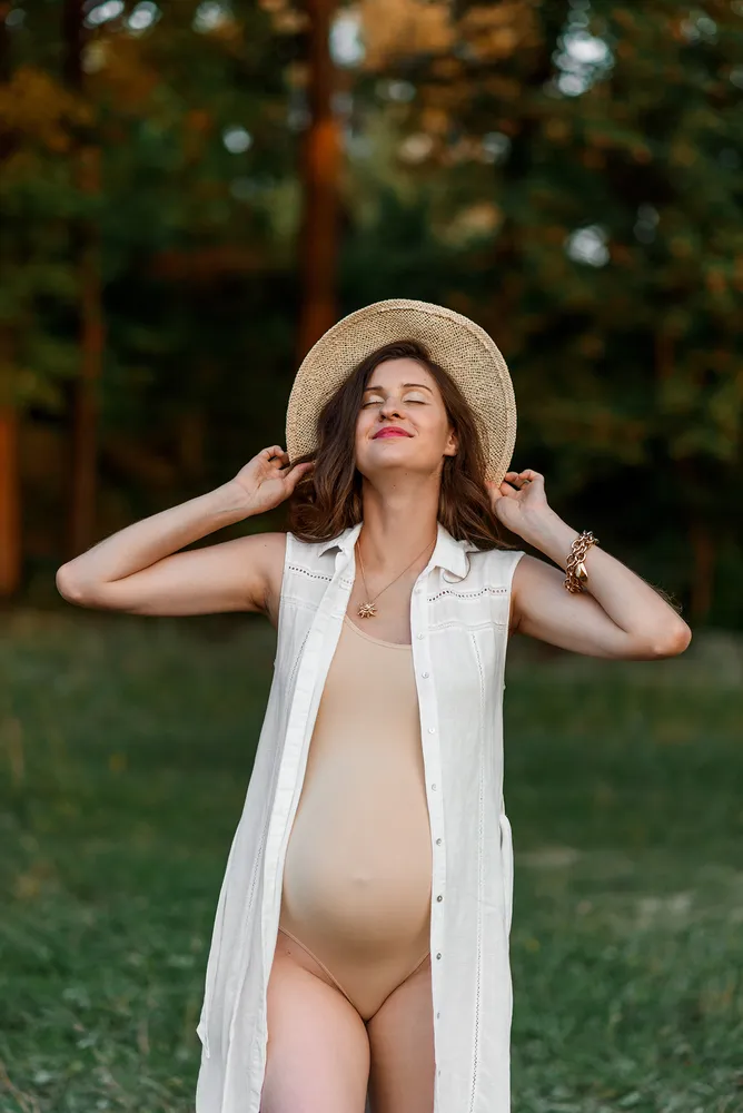 Woman in white shirt and straw hat adjusts hat in sunlit forest clearing.