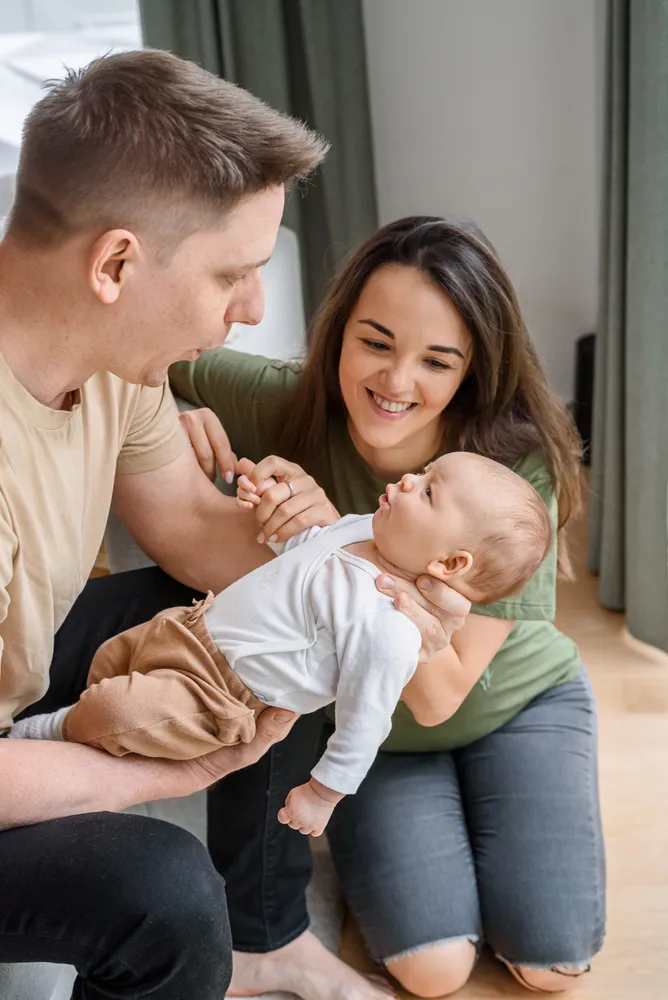Family bonding indoors: A man and woman joyfully interact with a baby on a wooden floor, surrounded by soft furnishings and curtains.