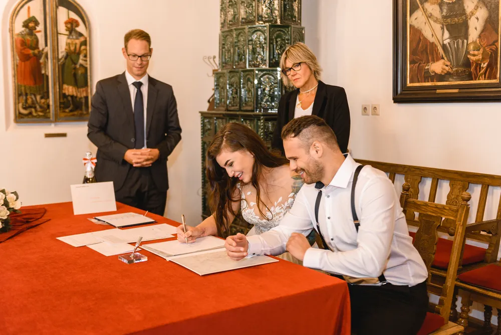 Bride and groom sign documents at a red-clothed table, observed by a formally dressed couple in a room adorned with historical artwork and a glass display case.