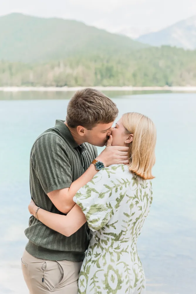 A couple shares a kiss by a serene lake, surrounded by a misty mountain landscape. The man wears a striped shirt and khaki pants; the woman is in a green and white patterned dress. Their affectionate embrace is captured in this intimate outdoor moment.