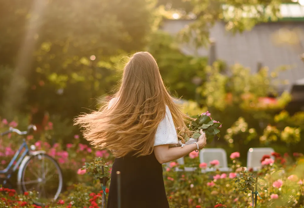 A woman with long blonde hair, dressed in a white blouse and black skirt, walks away from the camera in a lush garden, holding a bouquet of flowers. The background features a bicycle and a building, bathed in sunlight. She is adorned with a pearl bracelet, and the scene is filled with various shades of green and pink from the surrounding flora.