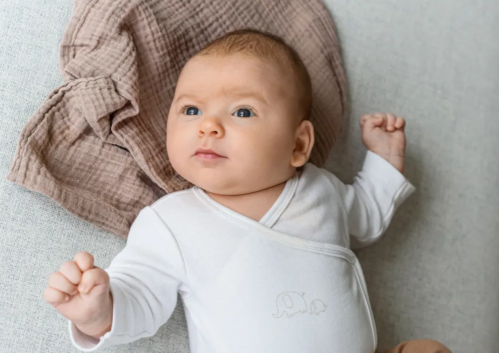 Infant in white onesie lies on light fabric, gazing upward. Brown textured cloth beside. Hands slightly raised, relaxed posture.