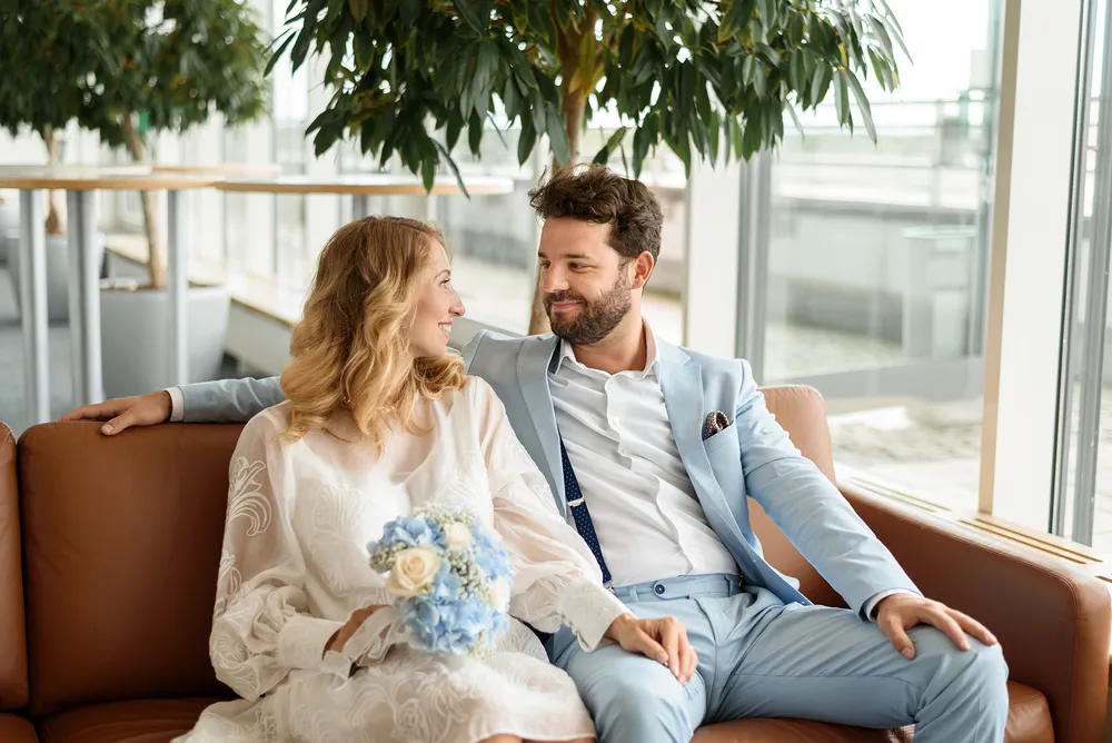 Couple in wedding attire sits on brown leather couch in modern office, holding bouquet, smiling at each other.
