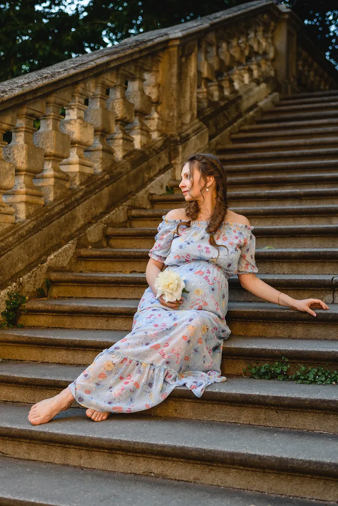 Woman in floral dress sits on stone steps, holding bouquet, gazing to left. Ornate balustrade, trees in background.