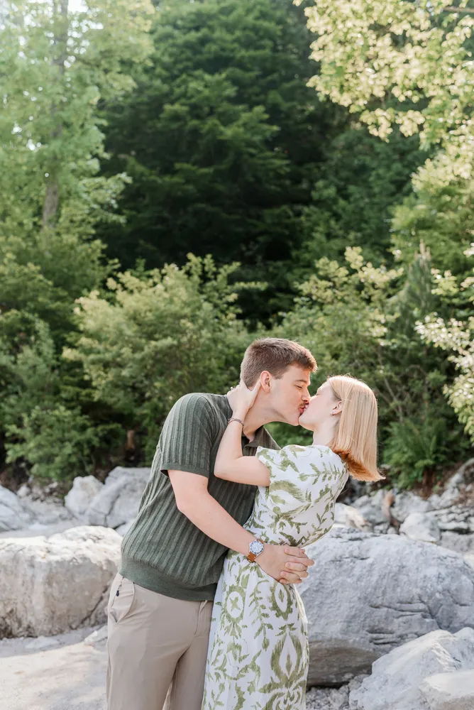 Couple sharing a kiss amidst a serene forest backdrop, surrounded by natural stone formations and lush greenery, capturing a moment of affection and tranquility.