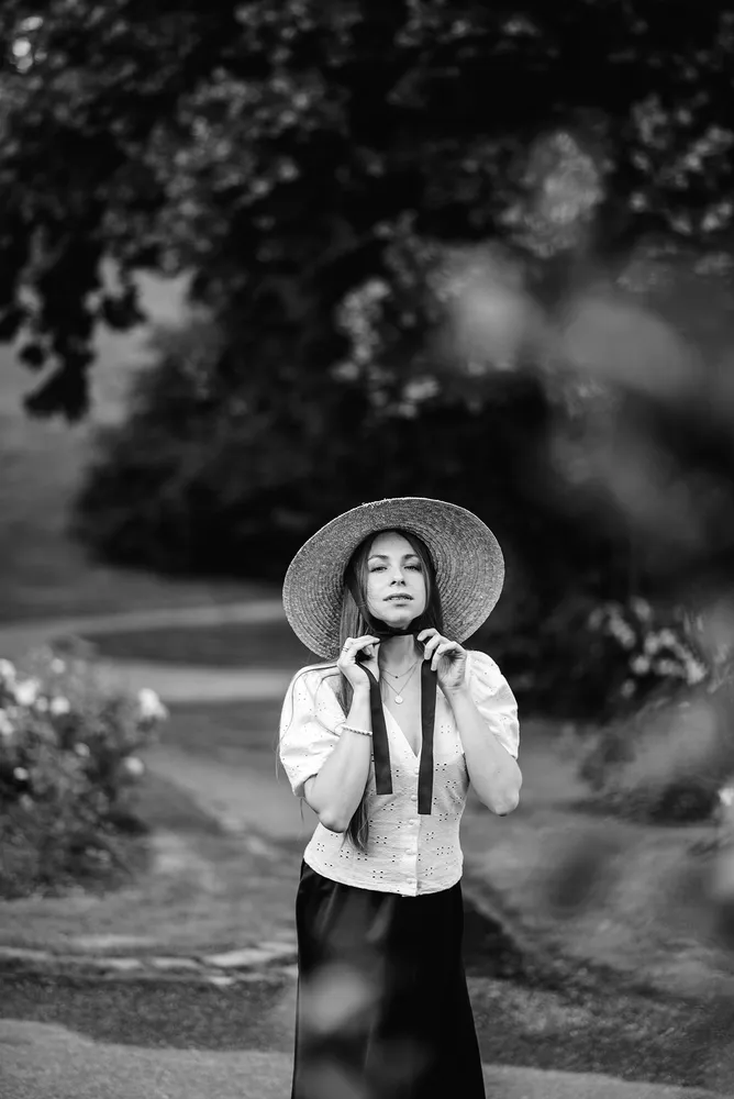 Woman in straw hat adjusts wide-brimmed hat outdoors, dressed in patterned blouse and dark skirt, surrounded by blurred foliage.