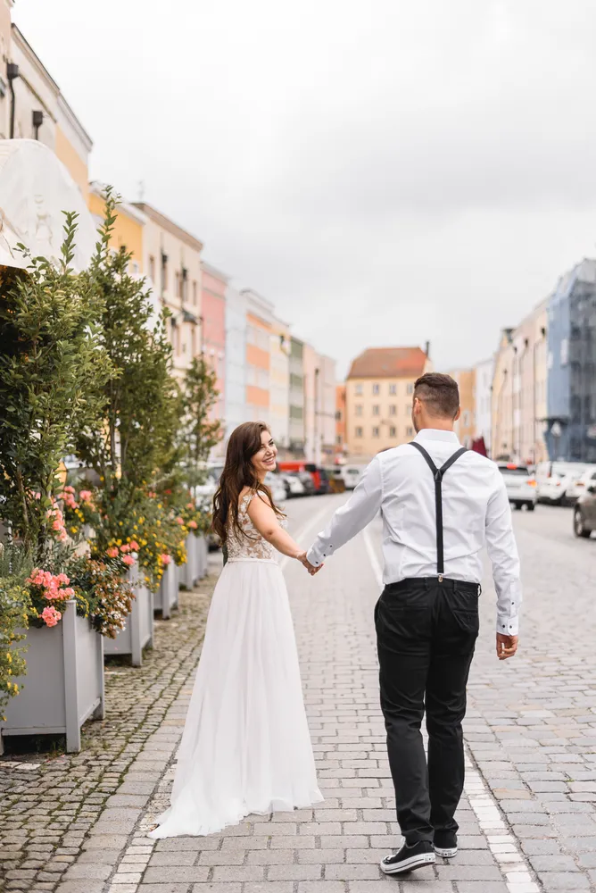 Couple strolling hand-in-hand on cobblestone street, woman in white lace dress, man in white shirt with suspenders, pastel buildings and blooming planters line the path.