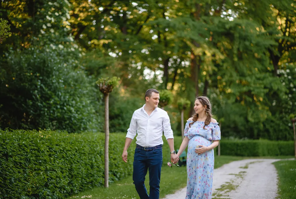 Expectant couple strolls through a lush park on a gravel path, holding hands and smiling. The man wears a white shirt and blue jeans; the woman is in a floral dress. Verdant trees and hedges frame the scene.