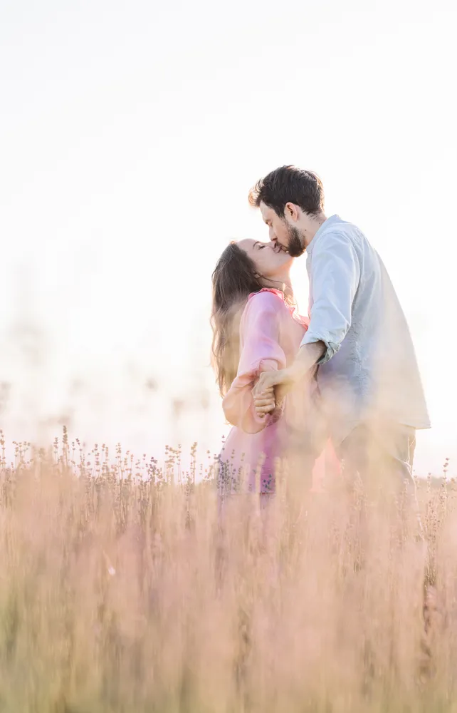Couple in a field, embracing and kissing, surrounded by tall, golden grasses. The woman wears a pink dress, the man a light blue shirt. Golden hour lighting creates a soft, romantic atmosphere.