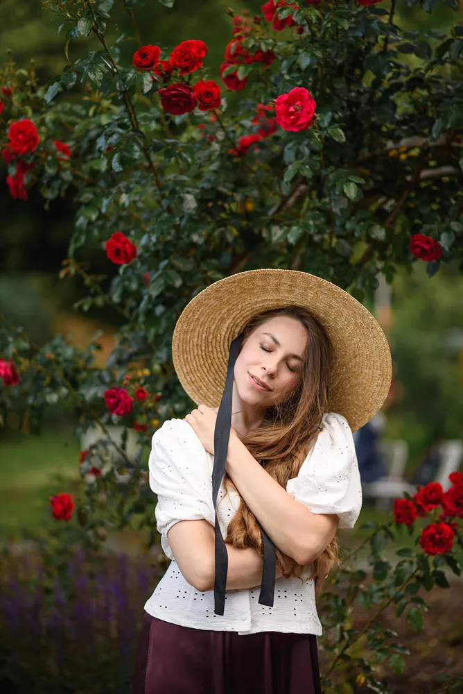 Woman in straw hat and white blouse stands amidst a rose garden, arms crossed, eyes closed in peaceful repose.