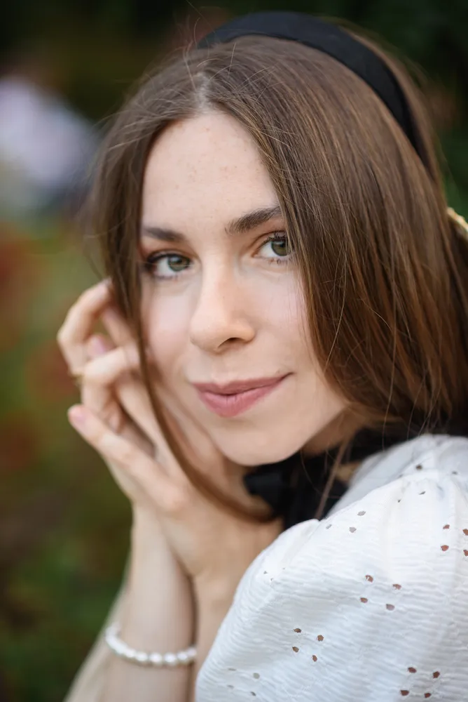 A woman with brown hair and green eyes wears a white lace top and pearl bracelet, resting her chin on her hands with a soft smile in an outdoor setting.