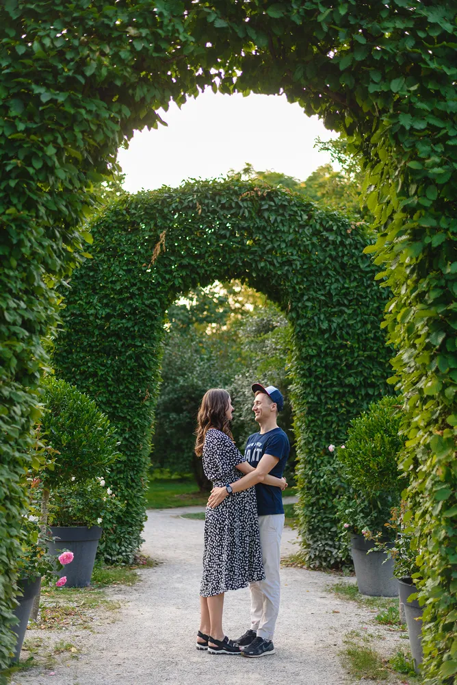 Couple in casual attire stands under a leafy garden archway, embracing. Man wears cap, woman in patterned dress. Gravel path, potted plants, and lush greenery surround them. Warm, golden-hour lighting enhances the romantic atmosphere.