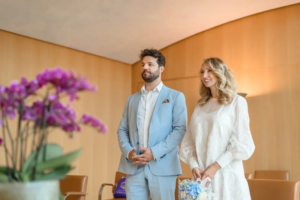 A couple in formal attire stands in a modern room with wooden paneling. The man wears a light blue suit, and the woman holds a bouquet, suggesting a wedding. They gaze off-camera, with a blurred vase of purple flowers in the foreground.