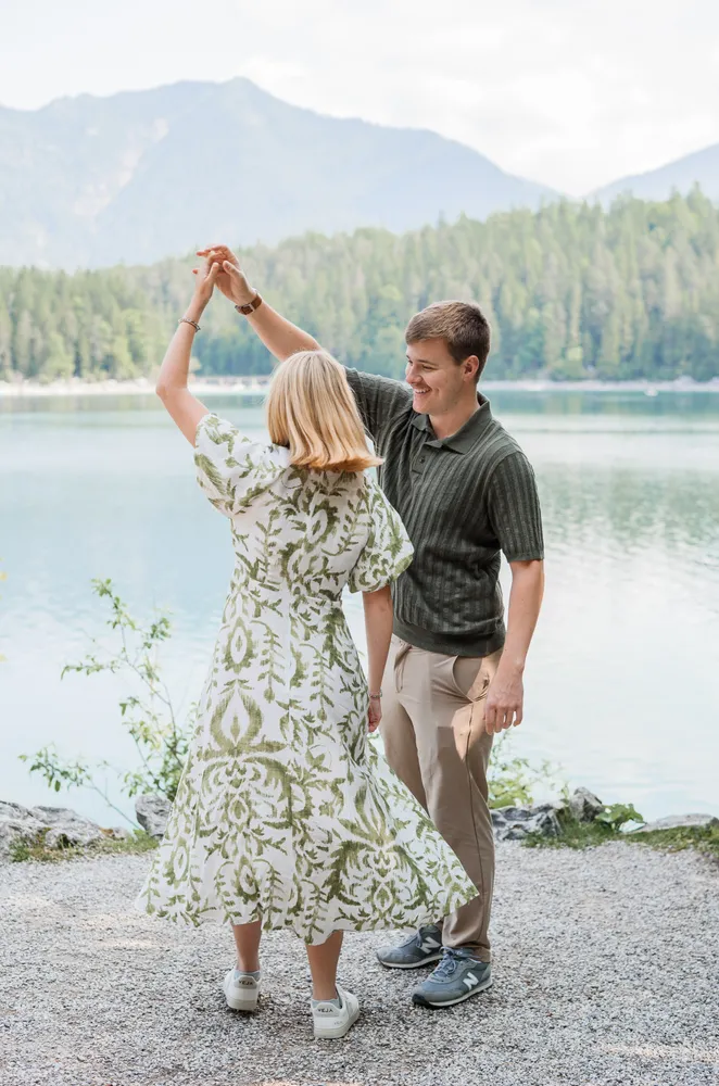 Couple dancing by serene lake, surrounded by mountains and trees. Woman in green floral dress, man in striped polo. Both smiling, joyful.