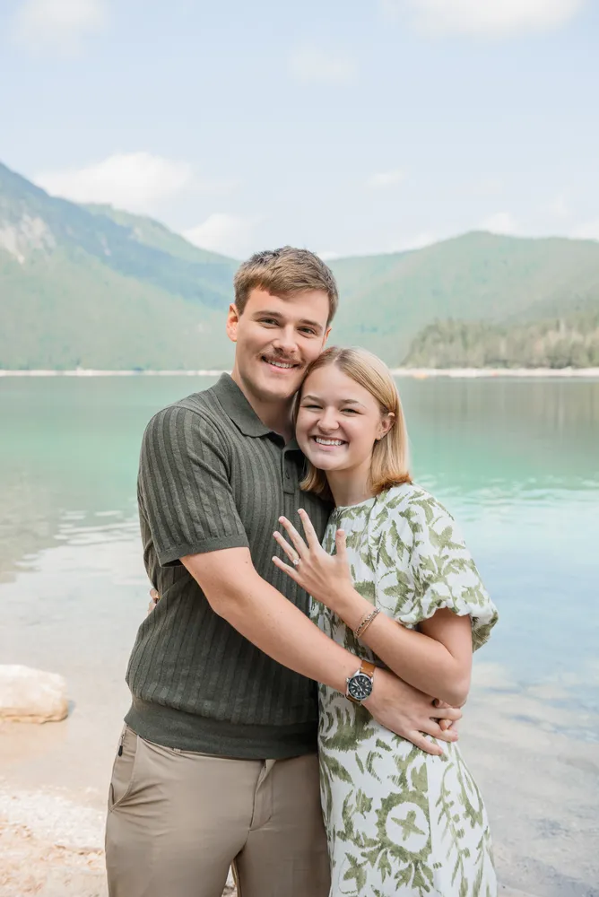 A couple shares a joyful embrace by a tranquil lake, surrounded by lush mountains. The man, wearing a striped polo and khaki pants, smiles broadly, while the woman, in a green floral dress, displays a ring on her left hand. Their affectionate pose and genuine smiles capture a moment of happiness in a picturesque natural setting.